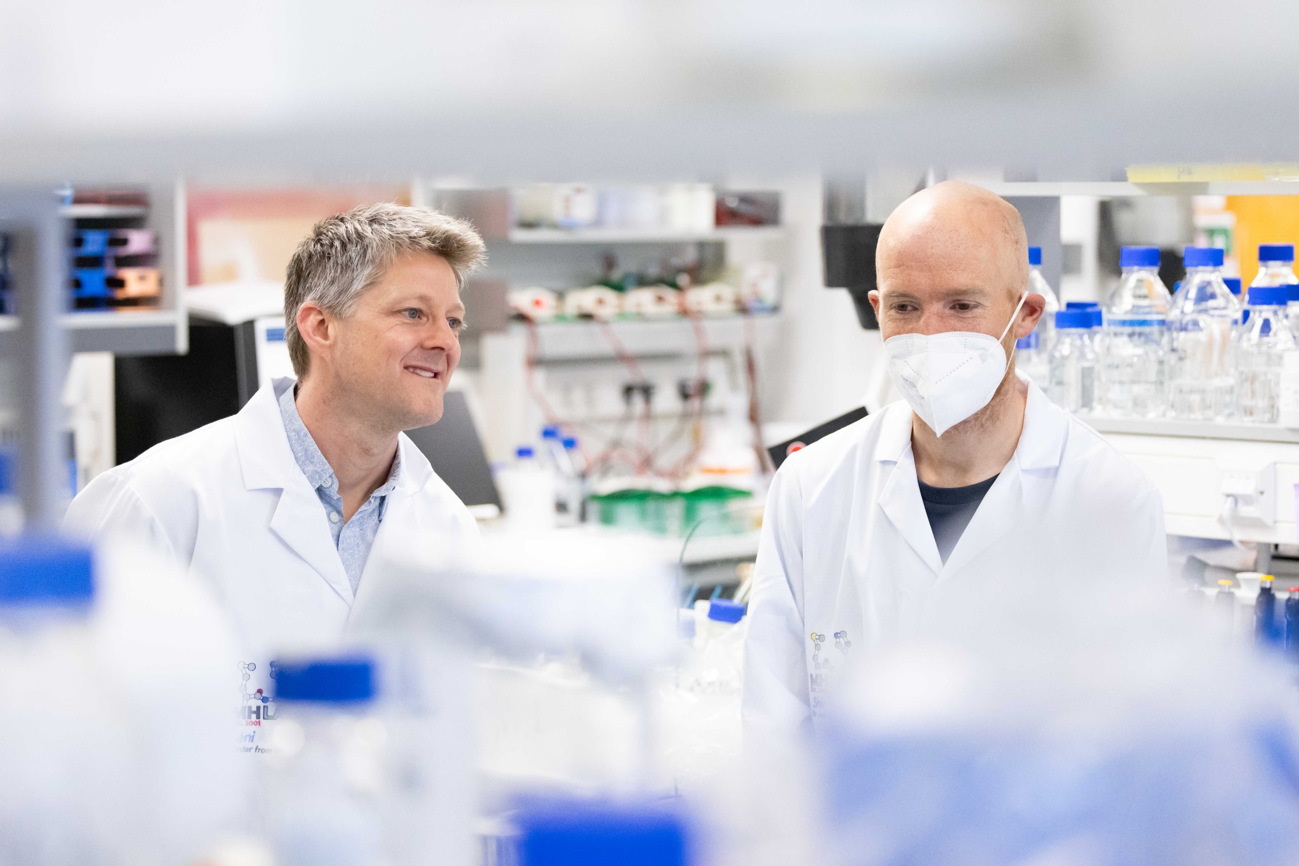 2 scientists in a lab with white coats on, one is wearing a mask. There are bottles and other lab items.