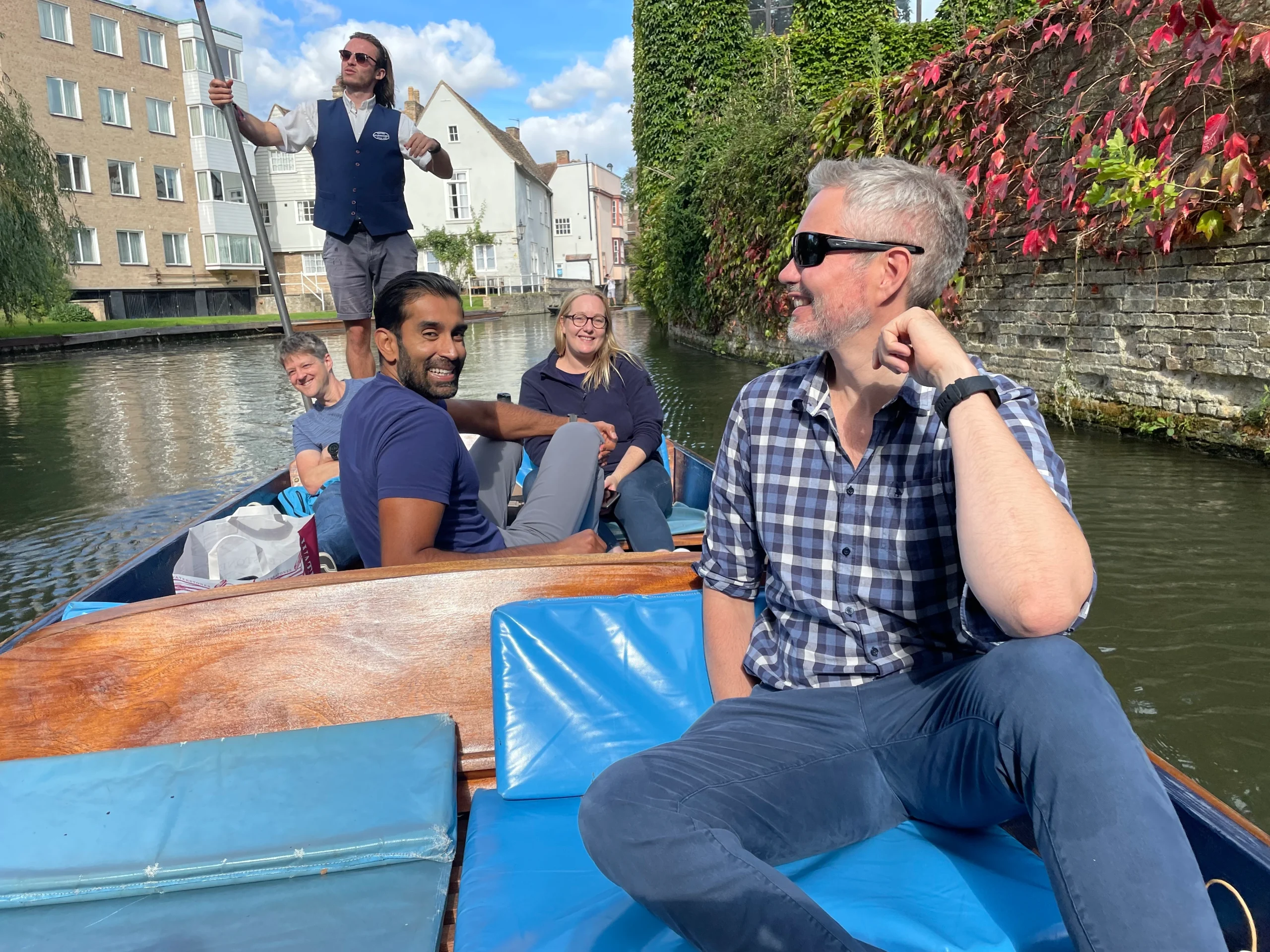 5 people on a punt in Cambridge looking relaxed. One person standing at the back with the punt pole.